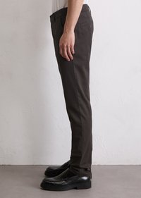 Side view of a person wearing dark brown slim-fit trousers and black leather loafers against a white textured wall.