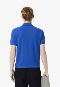 Nuori mies, jolla on lyhyet, aaltoilevat vaaleat hiukset, pukeutuneena siniseen polo-paitaan ja pitäen harmaata esinettä kädessään, seisoo valkoista taustaa vasten.