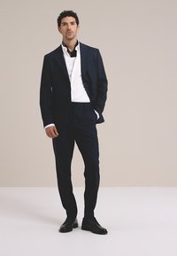 Donkerblauw marinepak met een getailleerde pasvorm, gecombineerd met een wit overhemd, afgewerkt met zwarte leren schoenen en een zwarte sjaal, tegen een neutrale achtergrond.