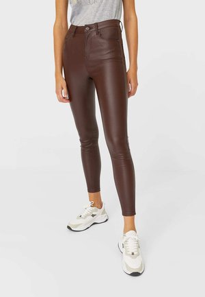 Person wearing brown faux leather skinny pants and white sneakers, standing against a plain light background.