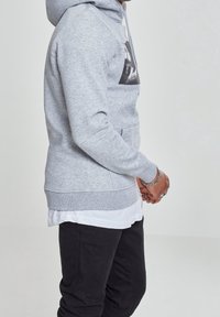 Felpa con cappuccio grigia in materiale misto cotone, con tasca frontale e motivo grafico sul petto. Abbinata a pantaloni neri e camicia bianca a strati.