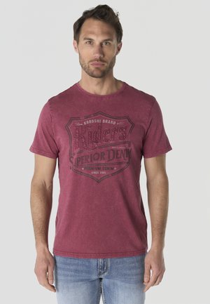 Hombre con cabello corto y castaño que lleva una camiseta gráfica roja con texto "Riders Superior Denim" y vaqueros azul claro, de pie frente a un fondo liso.