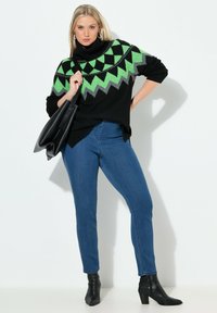 Mustat ylikokoiset neuleet, joissa on vihreä ja harmaa geometrinen kuvio, yhdistetty sinisiin farkkuihin ja mustiin nilkkureihin. Malli pitää mustaa laukkua.