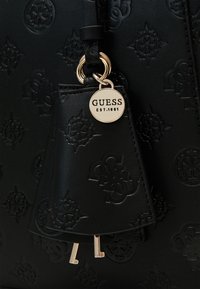 Musta nahkainen käsilaukku, jossa on kohokuvioitu logokuvio, kultainen etiketti kaiverrettuna teksti "GUESS EST.1981" ja metalliset avaimen yksityiskohdat.