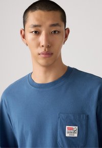 Blaues Baumwoll-T-Shirt mit kurzen Ärmeln, Rundhalsausschnitt und einer frontalen Tasche. Verfügt über ein Label mit den Markeninformationen "Levi's" und "Pixar".