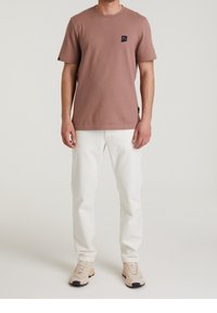 Mężczyzna w teksturowanej brązowej koszuli z krótkim rękawem, białych dżinsach i beżowych trampkach stoi na tle jasnym.