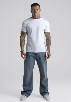 Mann mit Tattoos am Hals und an den Armen, der ein eng anliegendes weißes T-Shirt, lockere blaue Jeans und weiße Turnschuhe trägt und vor einem schlichten grauen Hintergrund steht.