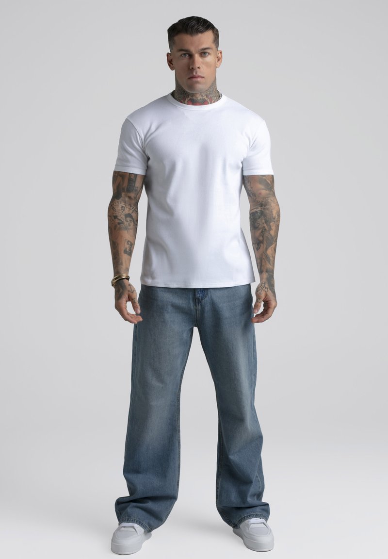 Homme avec des tatouages sur le cou et les bras portant un t-shirt blanc ajusté, un jean bleu ample, des baskets blanches, debout devant un fond gris uni.