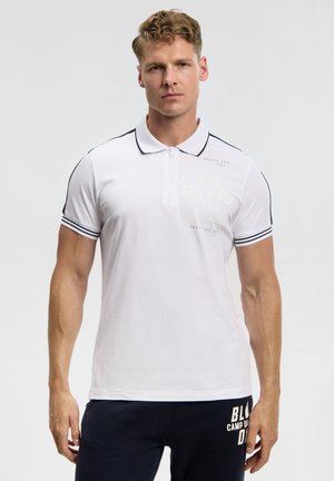 Mann trägt weißes, kurzärmeliges Poloshirt mit schwarzer Einfassung und marineblaue Hose mit weißem Text, steht vor schlichtem, hellem Hintergrund.