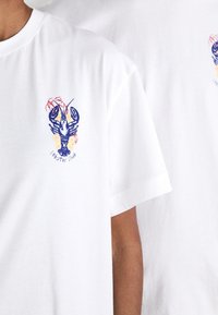 Bílé trička s malými modrými ilustracemi humrů a textem "lobster club" na levé části hrudi, které nosí dvě osoby.