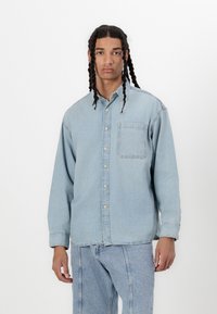 Cămașă din denim de un albastru deschis, cu închidere cu nasturi în față, guler și un buzunar pe piept. Croi lejer și mâneci lungi, cu o textură subtilă.