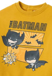 Geel sweatshirt met een Batman-graphic, twee cartoonvleermuizen en de tekst "THE BATMAN" en "ZAP" in vette, zwarte en oranje accenten.