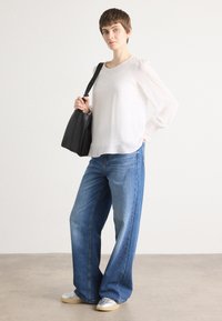 Lichte, doorzichtige blouse met lange mouwen in het wit, gecombineerd met losse blauwe denim jeans en zilveren sneakers. Model houdt een zwarte tote bag vast.