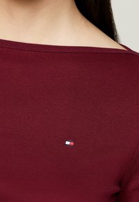 Top a maniche lunghe bordeaux con una texture a costine e un sottile logo con accenti rossi, bianchi e blu nella zona del petto sinistro.