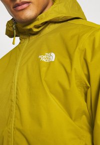 Chubasquero amarillo hecho de tela ligera con un patrón texturizado. Presenta un cuello alto, capucha ajustable y un logo blanco en el pecho.