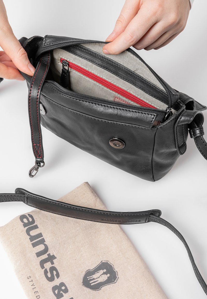 Zwarte leren crossbody tas met een ritsluiting, gestreepte voering, rode accenten en een buitensnapknop. Inclusief een stoffen beschermtas.