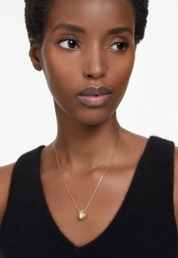 Collar de oro con un colgante de piedra redonda, colgado de una fina cadena. Lleva puesto sobre una blusa sin mangas negra y texturizada.