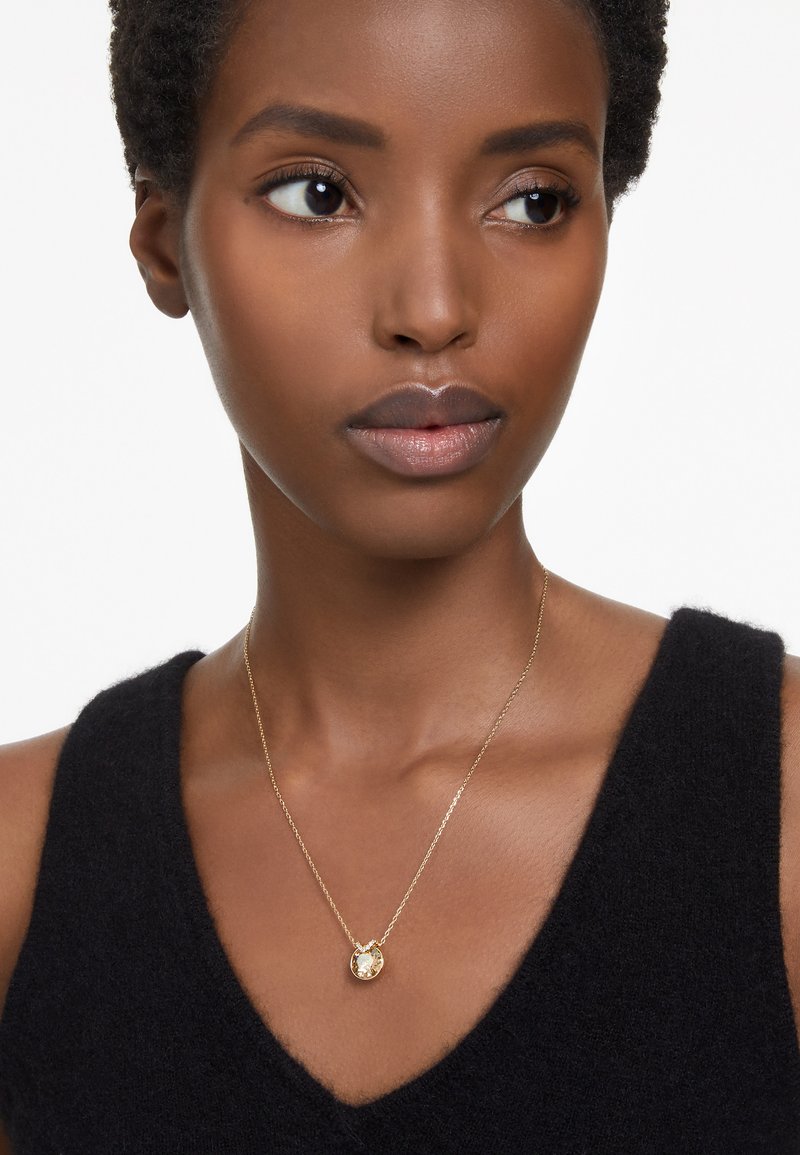 Collar de oro con un colgante de piedra redonda, colgado de una fina cadena. Lleva puesto sobre una blusa sin mangas negra y texturizada.