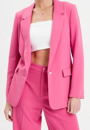Femme portant un costume rose vif avec un blazer et un pantalon taille haute, associé à un haut tube blanc court et des colliers dorés superposés.