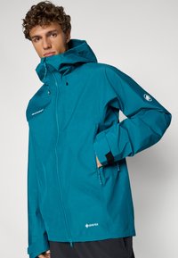 Teal waterproof jacket made of GORE-TEX, featuring a front zip, adjustable hood, and Velcro cuffs. Minimalist design with branding accents.