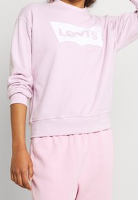 Sweat-shirt rose clair à col rond et poignets côtelés, avec un logo Levi's blanc sur le devant. Texture douce, coupe décontractée.