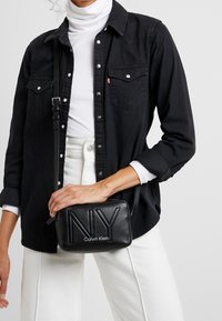 Borsa a tracolla in pelle nera con logo embossato "NY" e "Calvin Klein". Presenta una chiusura con zip e una tracolla regolabile. Texture liscia, forma rettangolare.