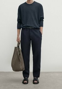 Maglione a maniche lunghe grigio scuro, pantaloni larghi blu, sandali neri e una borsa a tracolla in suede marrone. Terreno piatto e texturizzato.