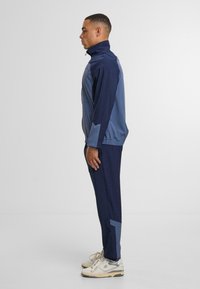 Navy un gaiši zils sporta treniņtērps ar rāvējslēdzēja jaku, pieguļošām biksēm, elastīgiem rokaviem un ģeometriskas krāsas bloku detaļām.