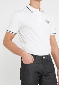 Bílá polo košile s černě vyšit�ým logem na hrudi, s dvoubarevnou légou a černě pruhovanými manžetami. V kombinaci s tmavě šedými kalhotami.