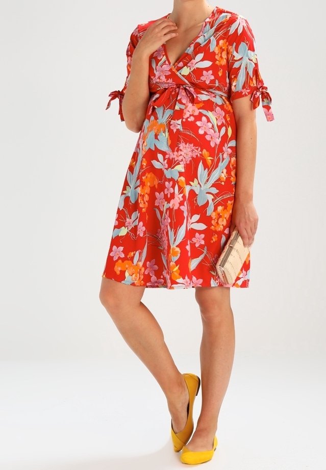 Femme en robe rouge fleurie longueur genoux tenant une pochette beige, portant des ballerines jaunes, posant devant un fond blanc uni.