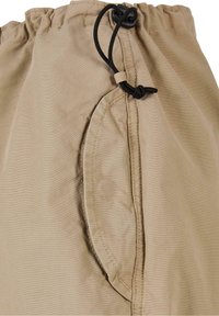 Tissu beige avec une taille élastiquée froncée et un cordon noir réglable noué en nœud près d'une poche latérale.
