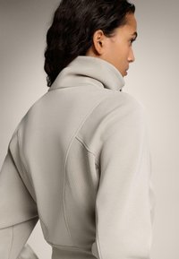 Svetlošedá mikina s vysokým golierom, rebrovaním na pleciach a hladkým textúrovaním látky, zobrazená zozadu.