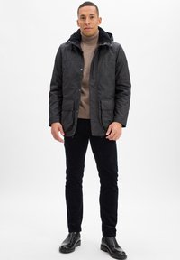 Mężczyzna w czarnej kurtce na brązowym golfie, czarnych spodniach i czarnych butach stojący na tle białym.