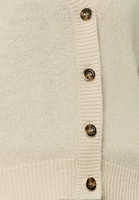 Kremowy kardigan z dzianiny, z ribbowym wykończeniem i rzędem sześciu ciemnych guzików w kolorze żółwia na przodzie. Gładka tekstura, casualowy styl.