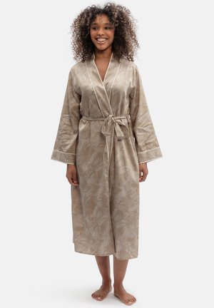 Femme aux cheveux bouclés souriante, portant une robe de chambre beige à motifs nouée à la taille, debout pieds nus devant un fond uni.