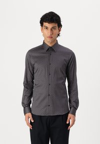 Mouwlange button-up shirt van donkere stof met een geometrisch patroon, voorzien van een puntige kraag en voorste knopen, gedragen met donkere broeken.