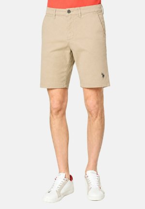 Uomo che indossa pantaloncini beige fino al ginocchio con tasche laterali e sneakers bianche, in piedi davanti a uno sfondo semplice.