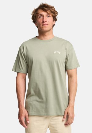 Man with wavy hair wearing a light green Billabong t-shirt and beige pants, standing against a plain white background.