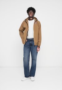 Brown lightweight jacket with yellow zipper, white t-shirt, distressed blue jeans, and white sneakers. Model stands against a white background.