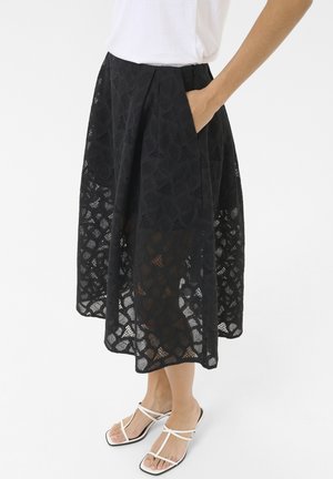 Jupe midi en dentelle noire présentant un motif ouvert texturé avec un design superposé et des poches latérales. Associée à des sandales blanches à brides.