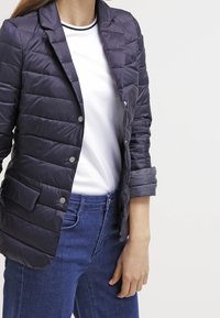 Kvinna som bär en marinblå vadderad blazer med rullade manschetter, en vit t-shirt med svart kant, och blå jeans mot en enfärgad vit bakgrund.