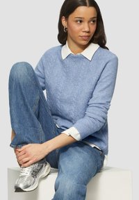 Blauwe gebreide trui met een gestructureerd ruitpatroon, gedragen over een wit overhemd met kraag. Gecombineerd met lichtblauwe jeans en zilveren sneakers.