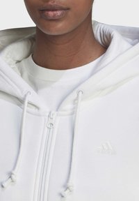 Veste à capuche blanche zippée en tissu doux, dotée de cordons de serrage réglables et d'un logo en relief subtil sur la poitrine.