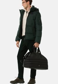 Mann in dunkelgrüner Steppjacke, cremefarbenem Pullover mit Reißverschluss am Kragen, dunkler Hose und Turnschuhen, der eine große schwarze, gesteppte Reisetasche hält.