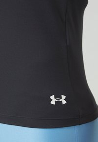 Gros plan sur une personne portant un haut noir Under Armour et un pantalon bleu clair, montrant le logo sur le côté inférieur du haut.