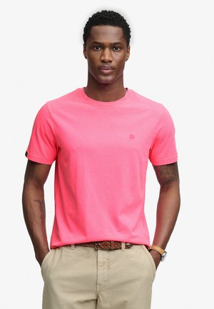Hombre con camiseta de manga corta rosa brillante, pantalones beige con cinturón marrón tejido y reloj de pulsera, de pie con las manos en los bolsillos.