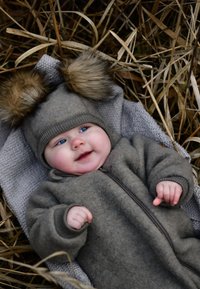 Siv volnen otroški komplet z bombicami iz krzna na kapici, z zadrgo in rebrastimi manšetami, položen na teksturirano sivo odejo s posušenim travjem.