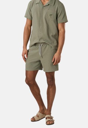 Man draagt een licht olijfgroen textuur kortmouwig overhemd met knopen en bijpassende short met trekkoord, gecombineerd met beige sandalen met gesp.