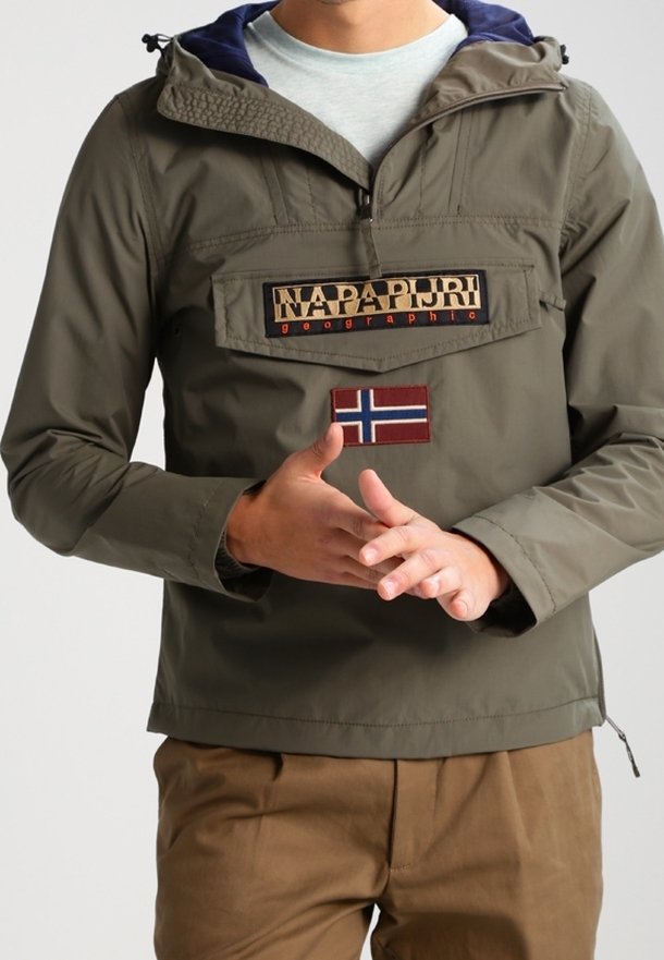 Uomo che indossa una giacca Napapijri verde oliva con tasca frontale e patch della bandiera norvegese, abbinata a pantaloni marroni.