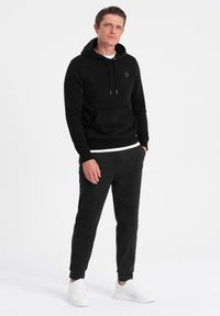 Schwarzer Hoodie mit Kängurutasche, Kordelzug und minimalem Logo. Kombiniert mit schwarzen Jogginghosen mit Paneeldetails und weißen Sneakers.